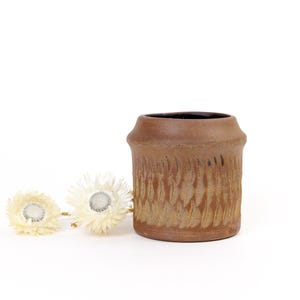 Könnte beinhalten: Eine kleine, braune Keramikvase mit einem strukturierten, wellenförmigen Muster. Die Vase ist leer und steht auf einer weißen Oberfläche.