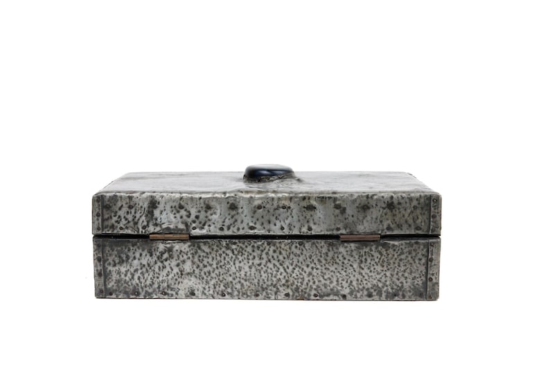 May include: A rectangular, silver-toned box with a textured surface and a black, rounded knob on the lid. The box has a hinged closure and appears to be made of metal. The overall aesthetic is rustic and industrial.