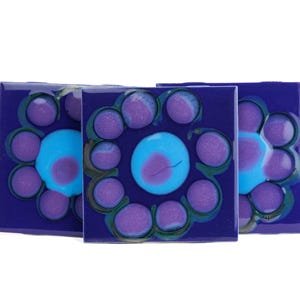 May include: Three square ceramic tiles with a floral design. Each tile has a deep blue background with a central light blue circle and surrounding purple petal-like shapes. The tiles are arranged in a slight offset.