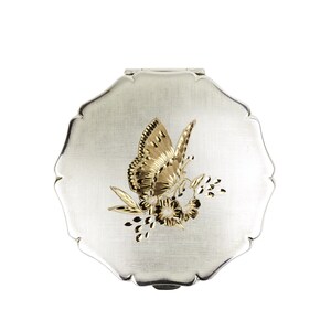 May include: A silver compact with a scalloped edge, featuring a gold butterfly and floral design. The butterfly is perched on a sprig of flowers, creating a decorative accent on the compact's surface.