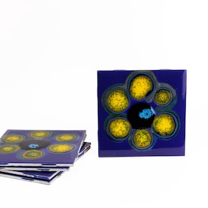 May include: A set of square ceramic coasters with a floral design. The coasters are a deep blue with yellow and black circular patterns. One coaster is standing upright, and a stack of coasters is to the left.