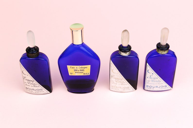 May include: Four blue glass bottles of perfume with silver and gold accents. The bottles are labeled "Evening in Paris" and "Soir de Paris" by Bourjois Paris.