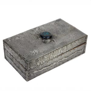 May include: A rectangular, riveted metal box with a textured, gray finish. The box has a hinged lid and a dark blue and green stone-like knob on top. The box is likely used for storage or display.
