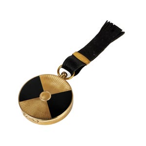 Könnte beinhalten: Ein goldfarbener Kompakt-Make-up-Spiegel mit einem schwarz-goldenen geometrischen Design. Der runde Kompaktspiegel hat einen schwarzen und goldenen Riemen mit Quaste. Das Kompakt ist ein Vintage-Accessoire.
