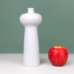 Puede incluir: Un jarrón de cerámica blanco con una parte superior abultada texturizada y un cuerpo cilíndrico alto. Una manzana roja se encuentra sobre una superficie blanca junto al jarrón.