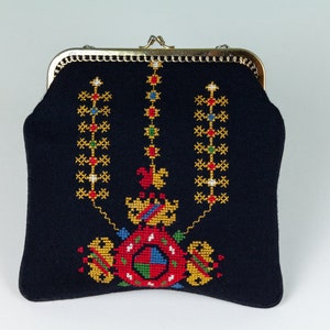 以下が含まれることがあります： 金色の金属製の留め具とカラフルな刺繍のデザインが施された黒い布製の財布。デザインは、赤、青、緑、黄色のアクセントのある中央のモチーフを特徴とし、その周りを3列の垂直な小さなモチーフが囲んでいます。