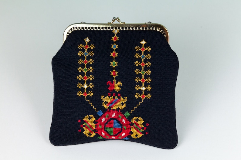 以下が含まれることがあります： 金色の金属製の留め具とカラフルな刺繍のデザインが施された黒い布製の財布。デザインは、赤、青、緑、黄色のアクセントのある中央のモチーフを特徴とし、その周りを3列の垂直な小さなモチーフが囲んでいます。
