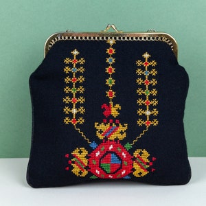 以下が含まれることがあります： 金色の金属製の留め具とカラフルな刺繍のデザインが施された黒い布製の財布。デザインは、赤、黄、緑、青の色で中央のモチーフを特徴としています。