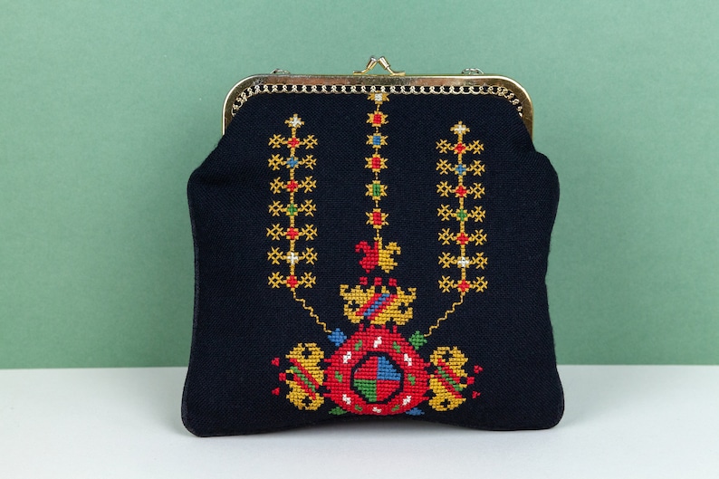 以下が含まれることがあります： 金色の金属製の留め具とカラフルな刺繍のデザインが施された黒い布製の財布。デザインは、赤、黄、緑、青の色で中央のモチーフを特徴としています。