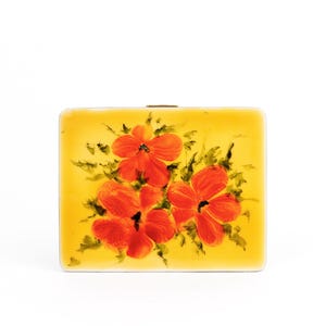 May include: A square, yellow compact case with a hand-painted floral design. The flowers are red with black centres and green leaves. The compact has a gold-coloured clasp.