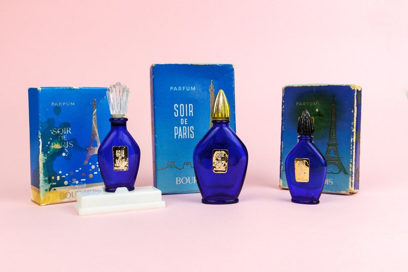 Puede incluir: Tres frascos de perfume Soir de Paris de vidrio azul con detalles dorados y tapones. Los frascos est&aacute;n sobre una superficie blanca con sus cajas originales detr&aacute;s. Las cajas son azules con una imagen de la Torre Eiffel y las palabras "Soir de Paris" en ellas.