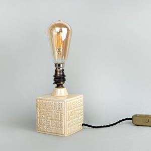 May include: A vintage table lamp featuring a cream-coloured, square base with a textured pattern. It has a large Edison bulb and a black cable with an on/off switch. The bulb emits a warm amber glow.