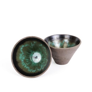 May include: A trio of cream-glazed ceramic mugs with a speckled finish. Each mug is adorned with a hand-painted design: a blue and green leaf, a yellow flower, and a red berry. The mugs have a simple, rounded form.