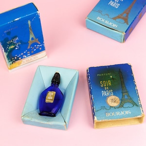Puede incluir: Tres cajas azules con letras doradas y una imagen de la Torre Eiffel. Las cajas est&aacute;n etiquetadas con "Soir de Paris" y "Bourjois". Un peque&ntilde;o frasco de perfume de vidrio azul con una tapa dorada se encuentra en una de las cajas.
