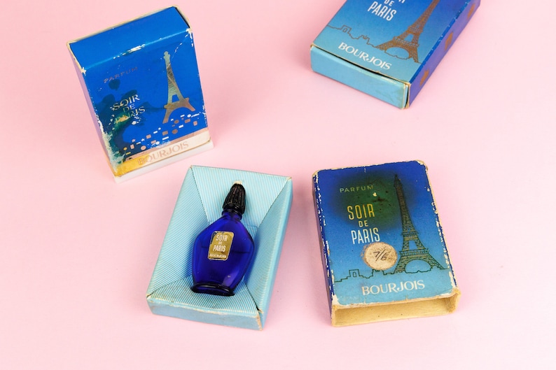 Puede incluir: Tres cajas azules con letras doradas y una imagen de la Torre Eiffel. Las cajas est&aacute;n etiquetadas con "Soir de Paris" y "Bourjois". Un peque&ntilde;o frasco de perfume de vidrio azul con una tapa dorada se encuentra en una de las cajas.