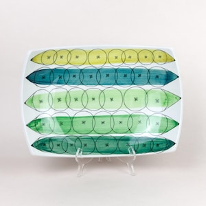 May include: Rectangular ceramic serving dish with a white background. The dish features four horizontal rows of colourful designs. Each row has a different colour: yellow, teal, light green, and dark green. The designs are made of overlapping circles and pointed ends.