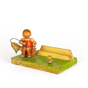 May include: A wooden figurine of a person sitting on a bench, holding a fishing rod with a fish on the line. The figurine is painted in bright colours, including red, yellow, and green. The bench is also painted in green and yellow. There is a small yellow duck figurine on the ground in front of the bench.