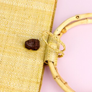 Puede incluir: Un bolso de paja tejido con un asa de bamb&uacute; natural. El bolso tiene un cierre de bot&oacute;n de madera marr&oacute;n.