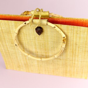 Puede incluir: Un bolso de mano de paja tejido con un asa circular de bamb&uacute;. El bolso es de color marr&oacute;n claro y tiene un forro naranja contrastante.