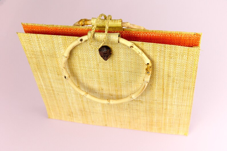 Puede incluir: Un bolso de mano de paja tejido con un asa circular de bamb&uacute;. El bolso es de color marr&oacute;n claro y tiene un forro naranja contrastante.