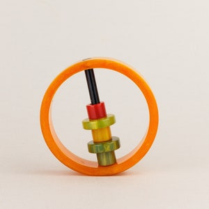 May include: A bright orange, circular toy with a black central rod and colourful, stacked rings. The rings are red, green, and yellow. The toy is set against a neutral background.