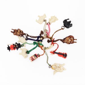 May include: A collection of small, colorful charms on cords, including animal and figure shapes. The charms are in various colors, such as brown, black, red, and white. The cords are in shades of brown, green, and pink.
