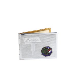 May include: Silver stamp holder with the word "Stamps" in a sign-like design. The holder has a textured pattern and a colorful emblem. The inside is lined with gold-colored material.