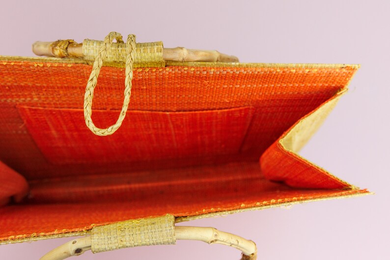 Puede incluir: Una bolsa de mano tejida de color beige y naranja con asas de bamb&uacute;. La bolsa est&aacute; abierta y se ve el interior. El interior de la bolsa est&aacute; forrado con tela naranja.