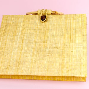 Puede incluir: Un bolso tote tejido de color beige claro con un asa de madera marr&oacute;n y un cierre de bot&oacute;n marr&oacute;n.