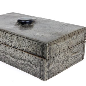 May include: A rectangular, riveted metal box with a textured, mottled gray finish. The lid features a small, rounded black stone as a handle. The box is likely used for storage and has a rustic, industrial aesthetic.