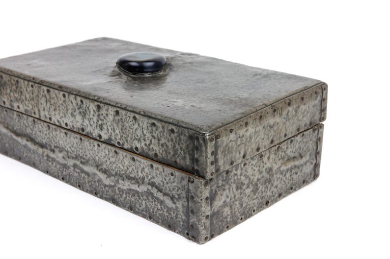 May include: A rectangular, riveted metal box with a textured, mottled gray finish. The lid features a small, rounded black stone as a handle. The box is likely used for storage and has a rustic, industrial aesthetic.