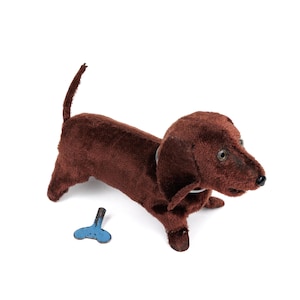 May include: A vintage wind-up toy dachshund dog in a rich brown colour. The toy has a long body, short legs, and a small tail. A blue wind-up key is next to the dog. The dog has a white collar.