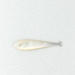 Könnte beinhalten: Ein dekoratives, fischförmiges Objekt mit Perlmutt-Effekt. Der Fisch hat einen hellen, schillernden Körper mit einem dezenten gelben Umriss und einem kleinen, dunklen Auge. Die Schwanzflosse ist grau. Das Objekt befindet sich vor einem schlichten, weißen Hintergrund.