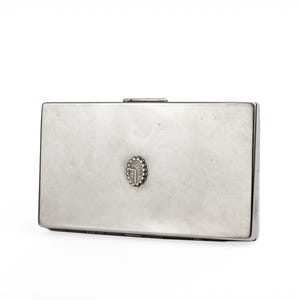 May include: A rectangular silver-toned metal box with a small, decorative emblem in the center. The box has a hinged lid and a simple, elegant design. The emblem appears to be a stylized design.