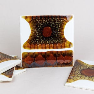 May include: A collection of ceramic tiles with a unique design. The tiles feature a white background with a central pattern in shades of brown, orange, and black. The design has a textured, organic appearance, with a central circular element.