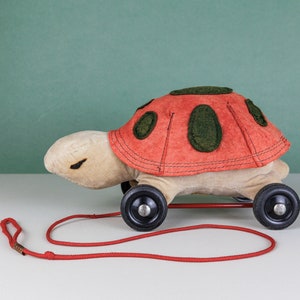 May include: A red and green toy turtle with black wheels and a red pull string. The turtle is made of felt and has a stitched design.