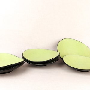 May include: Four light green, teardrop-shaped serving dishes with black rims and bases are arranged on a white background. The dishes are stacked in pairs and placed individually, showcasing their unique design.