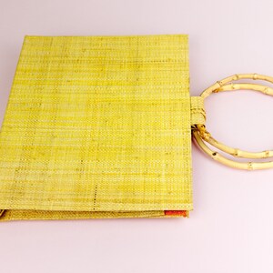 Puede incluir: Una bolsa de mano tejida de color amarillo con un asa de bamb&uacute; natural. La bolsa est&aacute; hecha de un material natural y tiene una textura tejida.