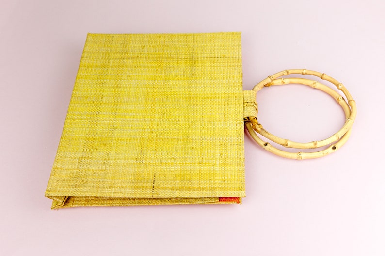 Puede incluir: Una bolsa de mano tejida de color amarillo con un asa de bamb&uacute; natural. La bolsa est&aacute; hecha de un material natural y tiene una textura tejida.