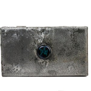 May include: A rectangular, metallic box with a textured, weathered appearance. A circular, dark blue and green decorative element is centered on the front. The box has a rustic, industrial aesthetic.