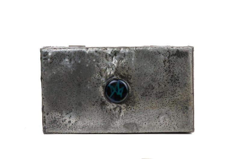 May include: A rectangular, metallic box with a textured, weathered appearance. A circular, dark blue and green decorative element is centered on the front. The box has a rustic, industrial aesthetic.