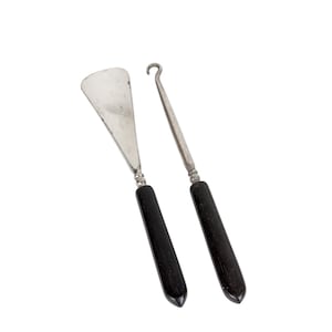 May include: Two antique shoe tools. One is a shoe horn with a silver blade and a black handle. The other is a long, slender tool with a hook at the end and a black handle. Both tools are set against a white background.