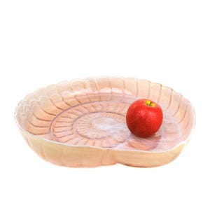 May include: A light-coloured, shell-shaped serving dish with a spiral design. A single red apple with a yellow stem rests inside the dish. The dish has a scalloped edge and is set against a white background.