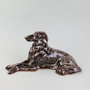 May include: A ceramic figurine of a brown and black dog lying down. The dog has a long, slender body and a long, pointed snout.