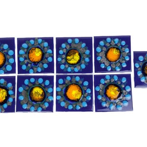 May include: A collection of nine square ceramic tiles. Each tile is a deep blue with a central design of a yellow-orange circle surrounded by gray and light blue accents. The tiles are arranged on a white surface.