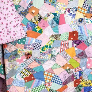 May include: A handmade quilt with a patchwork design. The quilt features a variety of colorful fabric scraps in geometric shapes, including pink, blue, yellow, and green. The quilt is folded over, revealing a pink floral patterned fabric.