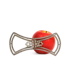 May include: A silver-toned bow-shaped brooch with clear, sparkling gemstones. The brooch is draped over a red apple, creating a striking contrast. The gemstones are set in a delicate, openwork design, adding elegance to the piece.