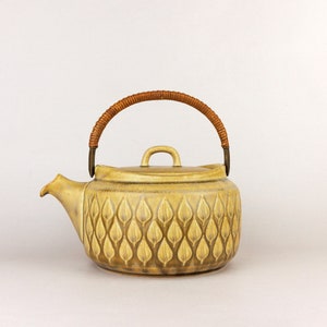 May include: A yellow ceramic teapot with a brown woven handle and a leaf pattern.