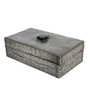 May include: A rectangular, gray metal box with a textured surface and a dark, oval-shaped stone on the lid. The box has visible rivets along the edges, suggesting a handcrafted design. The box is likely used for storage.