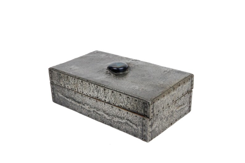 May include: A rectangular, gray metal box with a textured surface and a dark, oval-shaped stone on the lid. The box has visible rivets along the edges, suggesting a handcrafted design. The box is likely used for storage.